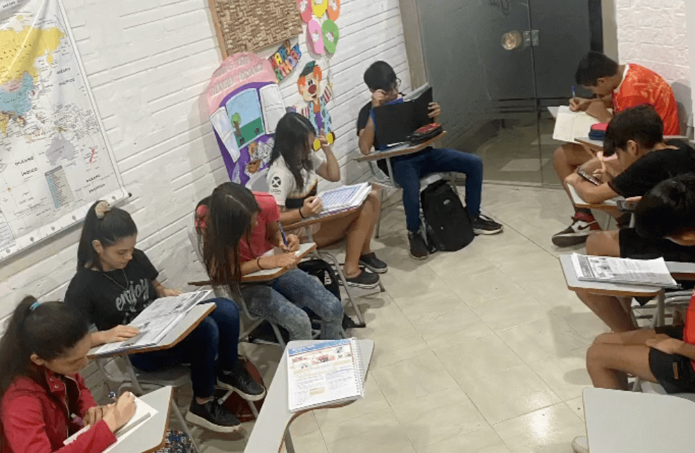 Alumnos en clase presencial de inglés, sentados frente a la docente al fondo del aula.